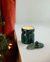 A green and blue striped candle next to a Christmas tree, enhancing the seasonal decor with its warm glow.