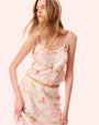 A woman wearing a floral dress and sandals stands gracefully, showcasing her vibrant outfit against a neutral background.