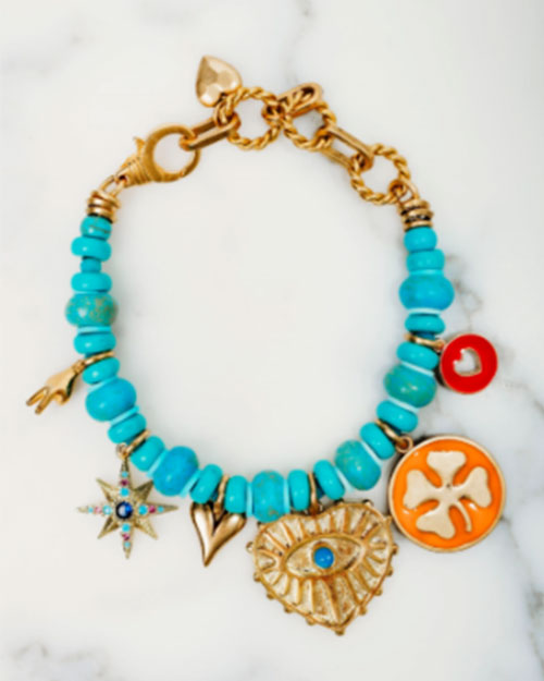A gold chain bracelet made up of turquoise beads with various colorful charms, in front of a marble background. 
