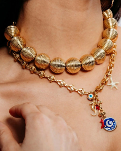 A model's neck wearing two gold necklaces. One necklace has a chain design with multiple small charms hanging from the middle. The second necklace has large, oversized, textured golden beads with vertical ribbing. 