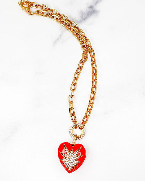 A gold chain necklace with a heart-shaped pendant in front of a marble background. The necklace has a starburst design in the center made of small white crystals, and the chain is linked to the pendant through a circular ring that is also adorned with crystals. 