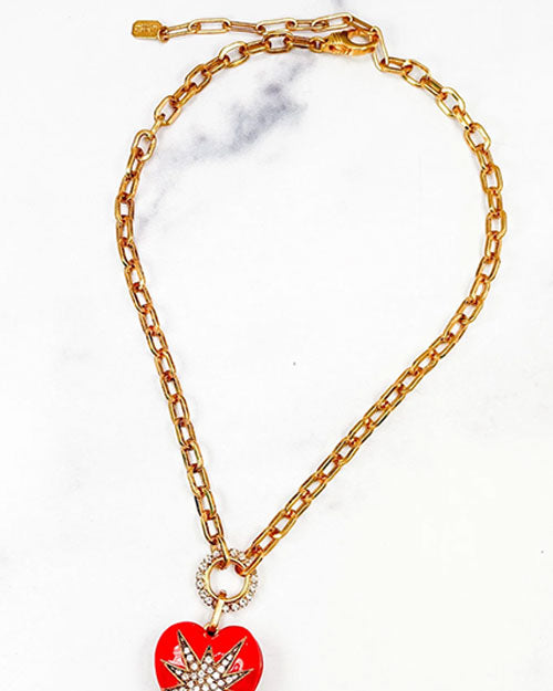 A close-up of a gold chain necklace in front of a marble background. The necklace features a starburst design in the center made of small white crystals. The chain is linked to the pendant through a circular ring that is also adorned with small white crystals. 