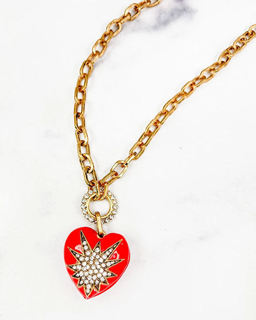 A gold chain necklace with a heart-shaped pendant. The pendant is red and features a starburst design in the center made of small white crystals. The chain is linked to the pendant through a circular ring that is also adorned with small white crystals. 