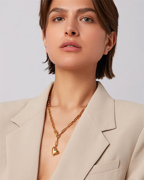 A woman wearing a beige blazer and a paperclip link chain necklace that has an inflated heart suspended from it stands elegantly, highlighting her fashionable look.
