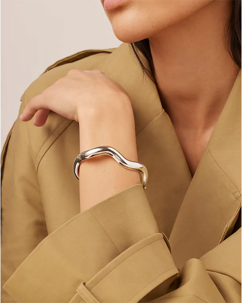 A woman dressed in a tan coat, showcasing a silver bracelet on her wrist