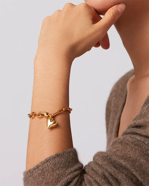 A woman showcases a gold bracelet adorned with a heart charm on her wrist.

