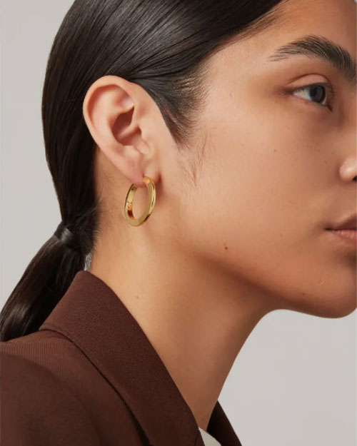 A woman in a brown jacket wears gold hoop earrings, showcasing a stylish accessory against her outfit.