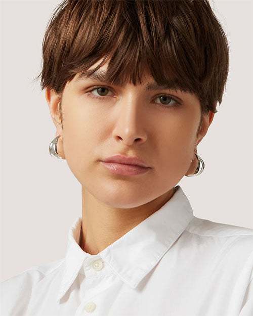 A woman with short hair is wearing a white shirt, looking confidently at the camera wearing a silver earring featuring a stylish curved design.