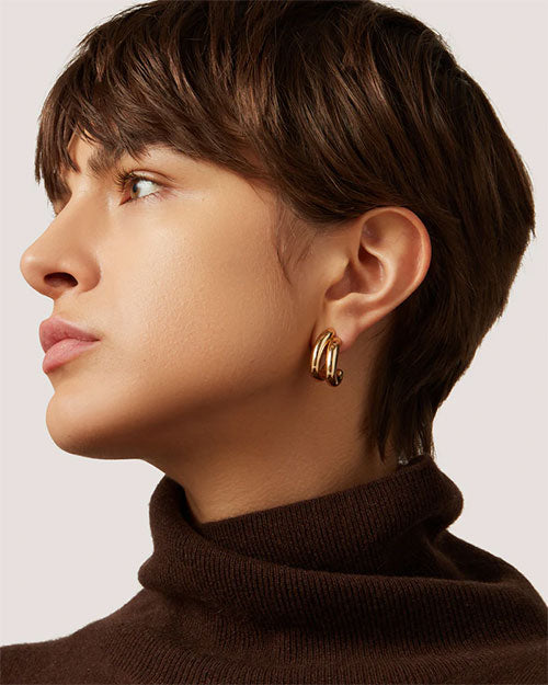 A woman in a brown turtleneck sweater and gold hoop earrings, smiling softly against a neutral background.
