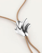A close-up image of a bolo tie, showcasing a unique seashell shaped silver pendant on a tan cord necklace. The pendant has an abstract, flowing design and is positioned at the center of the chest.