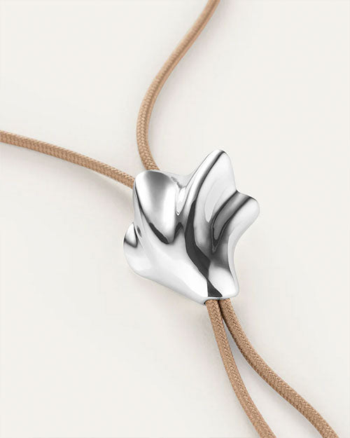 A close-up image of a bolo tie, showcasing a unique seashell shaped silver pendant on a tan cord necklace. The pendant has an abstract, flowing design and is positioned at the center of the chest.