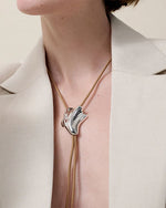 A person wearing a beige blazer with a deep neckline and a bolo tie, showcasing a unique seashell shaped silver pendant on a tan cord necklace. The pendant has an abstract, flowing design and is positioned at the center of the chest.