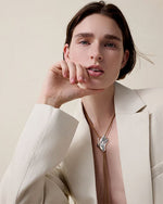 A person wearing a beige blazer with a deep neckline and a bolo tie, showcasing a unique seashell shaped silver pendant on a tan cord necklace. The pendant has an abstract, flowing design and is positioned at the center of the chest.