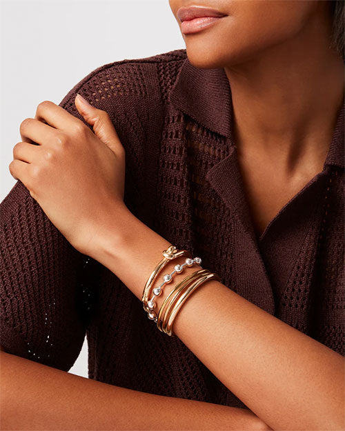 A model wearing a brown knit button-down top. She is wearing multiple gold bangles and a silver bracelet featuring small spherical beads evenly spaced around it.