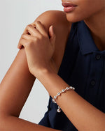 A model wearing a sleeveless, dark blue button-down top against a plain background. The model's right arm is crossed over her chest, and the person is wearing a silver bracelet with round beads evenly spaced around it and a dangling sphere charm.