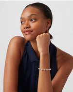A model wearing a sleeveless, dark blue button-down top. The model's jaw is resting on her hand, and she is wearing a silver bracelet with round beads evenly spaced around it.