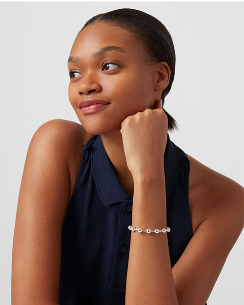 A model wearing a sleeveless, dark blue button-down top. The model's jaw is resting on her hand, and she is wearing a silver bracelet with round beads evenly spaced around it.