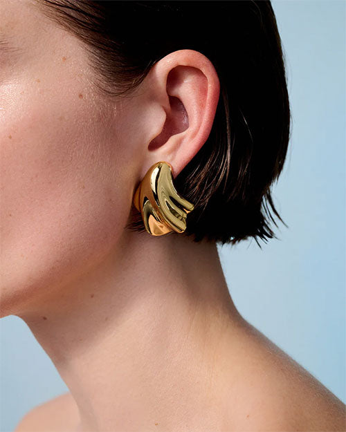 A close-up shot of the ear of a short-haired brunette model wearing a gold earring with an abstract, fluid design which resembles a seashell.