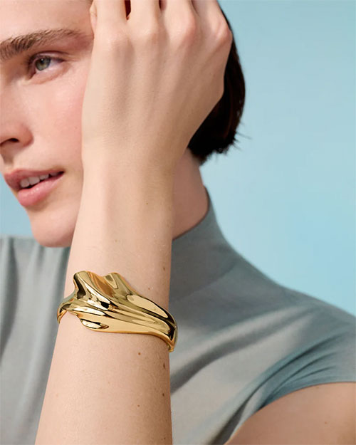 A person wearing a gold cuff bracelet on the wrist. The bracelet has an abstract, fluid design which resembles a seashell. The person is wearing a light grey top with short sleeves, and the background is a solid light blue color.