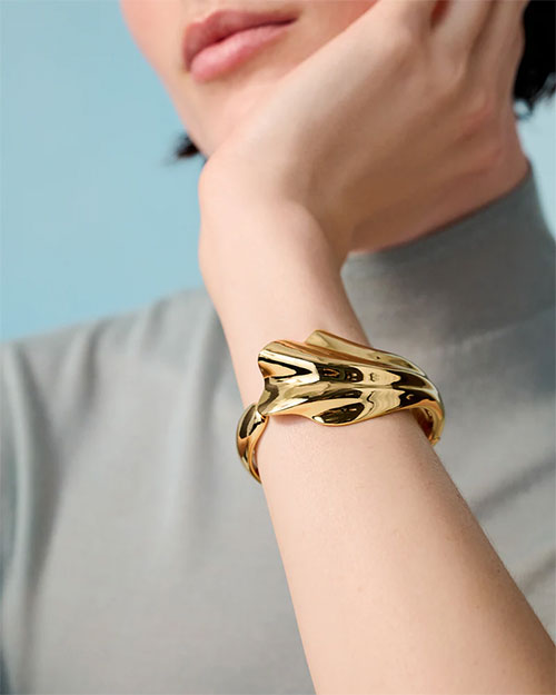 A person wearing a gold cuff bracelet on the wrist. The bracelet has an abstract, fluid design which resembles a seashell. The person is wearing a light grey top with short sleeves, and the background is a solid light blue color.
