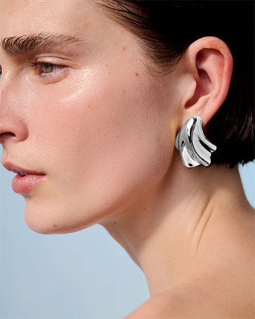 A sideview headshot of a model wearing a silver earring with an abstract, fluid design which resembles a seashell.