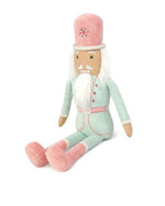 A plush toy nutcracker soldier sitting down in front of a white background. The toy has peppermint decorations, white hair and a beard and is dressed in a light teal/mint uniform with pink boots.