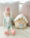 A plush toy nutcracker on a beige couch dressed in pastel mint pink, it is sitting next to a plush gingerbread house.