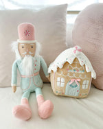 A plush toy nutcracker on a beige couch dressed in pastel mint pink, it is sitting next to a plush gingerbread house.