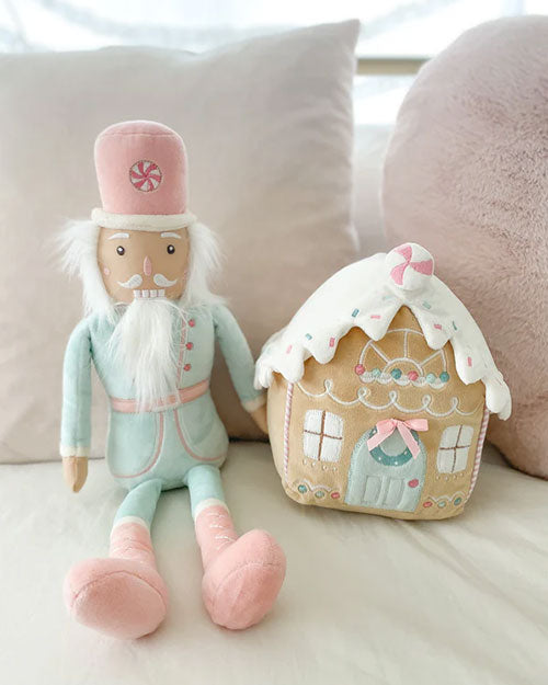 A plush toy nutcracker on a beige couch dressed in pastel mint pink, it is sitting next to a plush gingerbread house.