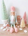 A pastel-colored Christmas scene with a nutcracker doll, a gingerbread man, and various festive decorations including colorful mini-Christmas trees, a gingerbread man, and a miniature gingerbread house. The nutcracker doll is dressed in a mint and pink uniform with a white beard and mustache, sitting on a wooden table. 