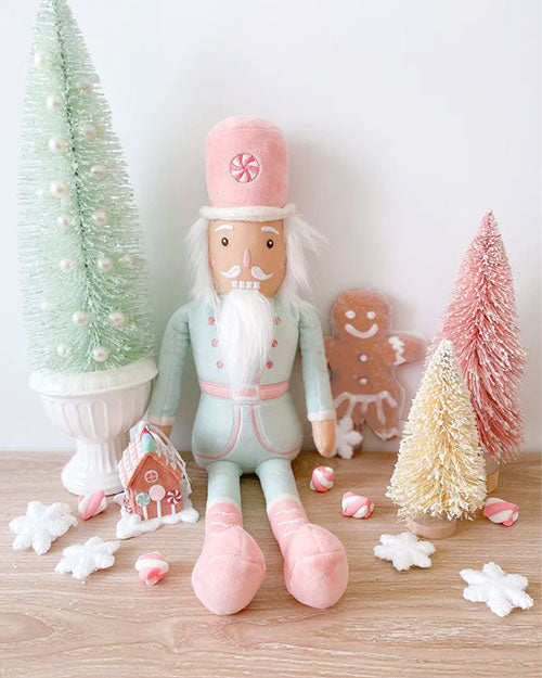 A pastel-colored Christmas scene with a nutcracker doll, a gingerbread man, and various festive decorations including colorful mini-Christmas trees, a gingerbread man, and a miniature gingerbread house. The nutcracker doll is dressed in a mint and pink uniform with a white beard and mustache, sitting on a wooden table. 