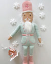A plush toy nutcracker laid on a white table, dressed in a pastel mint and pink uniform. The toy has a white beard, a pink hat with a peppermint candy design on it and is standing next to a small gingerbread house ornament. Surrounding the toy are five white snowflake decorations.