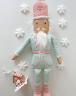 A plush toy nutcracker laid on a white table, dressed in a pastel mint and pink uniform. The toy has a white beard, a pink hat with a peppermint candy design on it and is standing next to a small gingerbread house ornament. Surrounding the toy are five white snowflake decorations.
