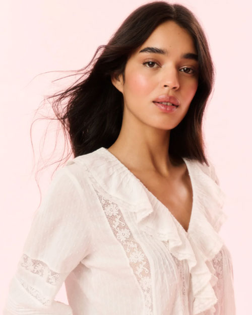 A close-up image of a brunette model wearing a white blouse with a ruffled neckline and a long neckline, including vertical lace detailing on the torso.