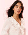 A close-up image of a brunette model wearing a white blouse with a ruffled neckline and a long neckline, including vertical lace detailing on the torso.