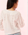 A waist-up image of the backside of a brunette model woman’s back is shown, wearing a white blouse with a ruffled neckline, vertical lace detailing along the torso, and horizontal detailing on the arms.