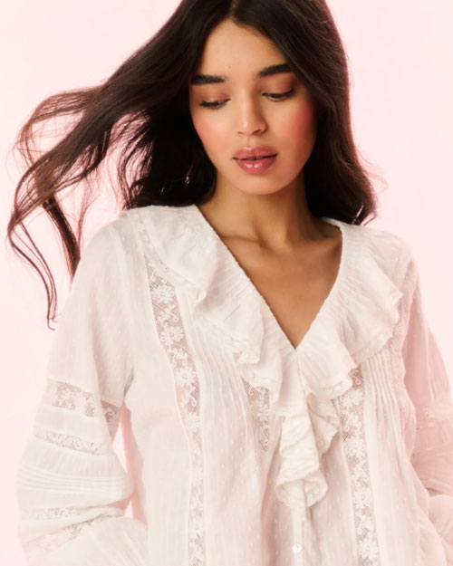 A close-up view of a brunette model with her hair blowing behind her, wearing a white blouse with a ruffled neckline, including vertical lace detailing on the torso and horizontal detailing across the arms.