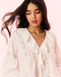A close-up view of a brunette model with her hair blowing behind her, wearing a white blouse with a ruffled neckline, including vertical lace detailing on the torso and horizontal detailing across the arms.