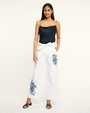 Woman wearing a navy top and white pants with blue floral embroidery on a white background