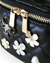 Close-up of a black quilted vanity-style handbag showing gold-tone zipper pulls and decorative floral embellishments in black, white, and crystal accents.