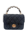 Small black denim handbag with a quilted diamond pattern, gold-tone twist-lock closure, and a short handle featuring interwoven black material and gold chain links, shown in a front view.