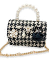 Small handbag with black and white houndstooth pattern, decorated with pearl and floral embellishments, a miniature black jacket ornament, and a black fabric bow. The handle is made of large pearls, and the bag has a gold chain strap.