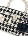 Detailed view of a black and white houndstooth handbag with pearl handle, showing decorative elements including pearl accents, a white enamel flower, a pearl cluster flower, and a miniature black jacket ornament with a black fabric bow.