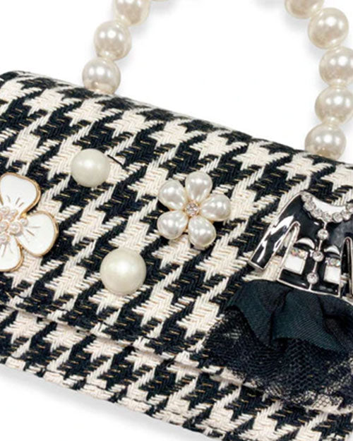 Detailed view of a black and white houndstooth handbag with pearl handle, showing decorative elements including pearl accents, a white enamel flower, a pearl cluster flower, and a miniature black jacket ornament with a black fabric bow.