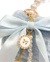 Close-up of a light blue faux fur handbag featuring a large satin bow, a gold ring attachment, and a decorative charm with pearls and a round white medallion accented with a small gold bow design.