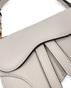 Close-up of a light gray saddle-style handbag showing the textured curved flap design and part of the patterned scarf tied to the handle.