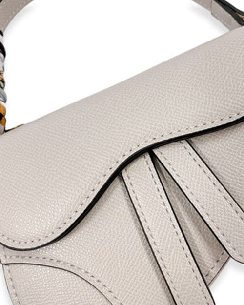 Close-up of a light gray saddle-style handbag showing the textured curved flap design and part of the patterned scarf tied to the handle.