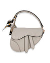 Small light gray saddle-style handbag with a curved flap design, accented by a patterned scarf tied around the short handle.