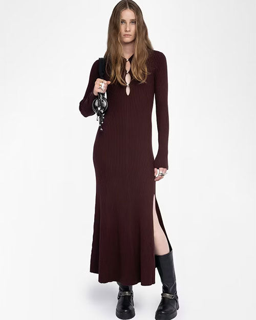 A person wearing a long, ribbed, dark burgundy knit dress with long sleeves and a front keyhole cutout detail. The dress has a side slit that reaches above the knee. The outfit is paired with chunky black boots featuring silver hardware and a black handbag with a circular metal handle. The person is accessorized with multiple rings.