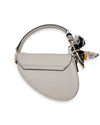 Back view of a light gray saddle-style handbag with a curved flap design and a patterned scarf tied around the short handle.